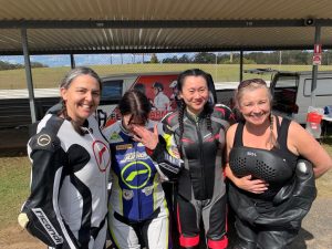 Four women in motorcycle gear smiling at a racing event